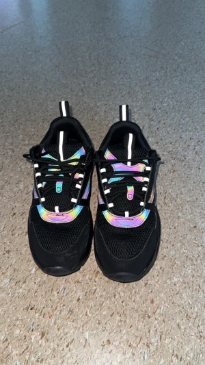 Svarta chunky sneakers med regnbågsdetaljer - Säljer ett par svarta chunky sneakers med mesh och syntetmaterial. Skorna har coola regnbågsfärgade detaljer som skiftar i ljuset och svarta snören med vita inslag. Snygg, sportig vibe och perfekt för dig som gillar att sticka ut med dina skor.