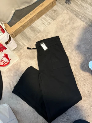 Svarta mjukisbyxor med snörning - Svarta mjukisbyxor med bred resår i midjan och snörning för extra komfort. Byxorna har raka ben och en stor ficka på sidan, perfekta för en chill och avslappnad stil.