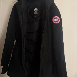 Svart dunjacka från Canada Goose - Säljer en svart dunjacka från Canada Goose med klassisk patch på ärmen och fluffig beige päls runt huvan. Jackan har dragkedja framtill och är långärmad, perfekt för kalla vinterdagar. Snygg och funktionell design med värmande material.