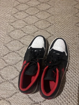 Nike Air Jordan 1 Low svart/röd - Nike Air Jordan 1 Low sneakers med svart och vitt skinn, röda detaljer och röd swoosh. Låg modell med perforerad vit tå, svart snörning och röd insida. Ikonisk vinge-logga på hälen och klassisk platt sula. Perfekt för dig som gillar streetstyle och sneakers med attityd.