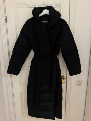 Svart lång pufferjacka med huva - Säljer en svart lång pufferjacka med huva och bälte i midjan. Jackan har knappar framtill och är quiltad för extra värme. Perfekt för kalla dagar och har en clean, oversized look som är riktigt chill.