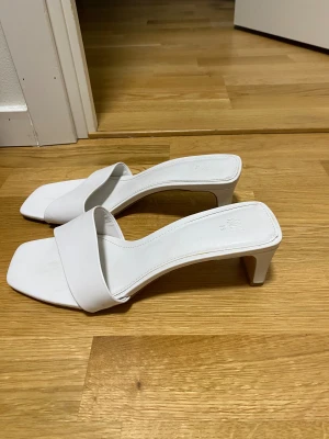 Vita klackade sandaler från H&M - Snygga vita sandaler från H&M med fyrkantig tå och bred rem över foten. Skorna har en låg klack och är tillverkade i syntetmaterial. Perfekta för dig som gillar stilrena och trendiga skor med minimalistisk vibe.
