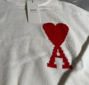 Vit stickad tröja från Ami - Vit stickad tröja från Ami med röd hjärtformad A-logga på bröstet. Tröjan har ribbad rund hals, muddar och nederkant. Materialet är mjuk bomull och passformen är normal. Perfekt för dig som gillar stilrena detaljer med en twist.