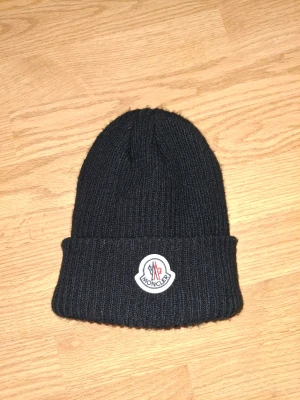 Svart Moncler mössa - Svart ribbstickad mössa från Moncler med klassisk uppvikt kant och broderad logotyp framtill. Tillverkad i mjuk ull som håller dig varm och ger en clean look. Perfekt accessoar för kalla dagar och snygg till streetwear.