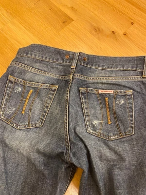 Blå bootcut jeans från Fornarina - Snygga blå jeans från Fornarina med klassisk low bootcut och slitna detaljer. Jeansen har fem fickor, gul kontrastsöm och knappstängning fram. Materialet är jeans med en tvättad look och lätt slitning vid benslut och fickor. Runt midjan är det 85cm. Länden på byxorna är 75cm. Runt låren är det 51-52cm.
