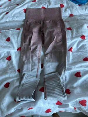 Rosa och grå ribbade leggings XS - Säljer ett par snygga leggings i ribbad kvalitet med hög midja. Färgen går i en cool ombre från rosa upptill till ljusgrå nedtill. Materialet är stretchigt med 65% nylon, 25% polyester och 10% spandex. Passar perfekt för träning eller chill. Scrunch-detalj bak för extra shaping.