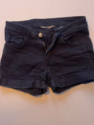 Svarta shorts från H&M med dragkedja - Säljer ett par svarta shorts från H&M med klassisk femficksdesign, uppvikta benslut och snygg dragkedja framtill. De har en metallknapp i midjan och är gjorda i mjuk bomull med lite stretch för skön passform.