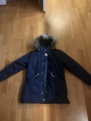 Mörkblå parkajacka med fuskpäls från Lindex - Parkajacka i mörkblå färg från Lindex med huva och fuskpäls. Jackan har dragkedja och knappar framtill, vita snören vid fickorna och är vattenavvisande. Perfekt för kalla dagar med sin långa modell och fodrade insida.