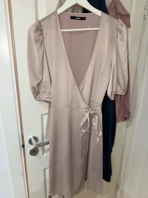 Ljus omlottklänning från Bik Bok - Säljer en ljusbeige omlottklänning från Bik Bok i storlek M. Klänningen har puffiga korta ärmar, v-ringad hals och knytband i midjan. Materialet är mjukt och har en lätt glansig yta som ger en elegant känsla. Perfekt för dig som gillar stilrena och feminina plagg.