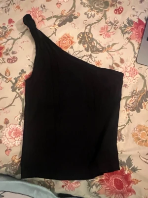 Svart oneshoulder topp - Säljer en svart oneshoulder topp med en snygg twistad axel. Toppen är ärmlös och har en stilren, tight passform som framhäver siluetten. Perfekt att matcha med jeans eller kjol för en cool look. Materialet känns mjukt och stretchigt mot huden.