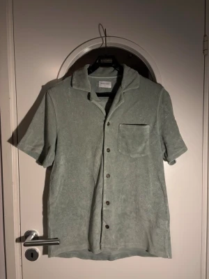 Ljusgrön frottéskjorta Studio Total - Snygg ljusgrön skjorta från Studio Total i mjuk frotté. Skjortan har korta ärmar, klassisk krage, bröstficka och knappar framtill. Perfekt för chill dagar och har en relaxed vibe. Passar dig som gillar stilren och enkel design.