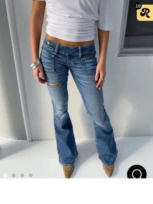 Blå bootcut jeans med slitningar - Säljer ett par blå bootcut jeans med låg midja och slitna detaljer vid knäna. Jeansen har framfickor med markerade sömmar och dubbla knappar i midjan. Materialet är klassisk jeans och passformen är loose med utställda ben.