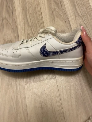 Nike Air Force 1 med blå detalj - Säljer ett par vita Nike Air Force 1 sneakers med blå sula och blå paisleymönstrad swoosh. Skosnöret till andra skin saknas och de är lite smutsiga vilket jag kan tvätta bort innan köp. 