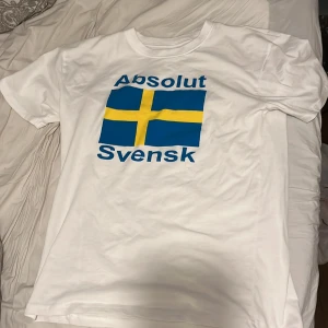 Vit t-shirt med svensk flagga - Vit t-shirt med tryck av den svenska flaggan och texten 'Absolut Svensk' i blått. Perfekt för att visa din svenska stolthet. Kortärmad och i bomull för en bekväm passform. OBS! Den är mindre än vad den ser ut nog mer en Medium