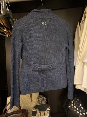 Blå stickad kofta från Odd Molly - Säljer en blå stickad kofta från Odd Molly med knytband i midjan och dekorativ detalj bak. Koftan har lång ärm, strukturstickat material och en liten etikett framtill. Perfekt för dig som vill ha en mysig och snygg vardagskofta.