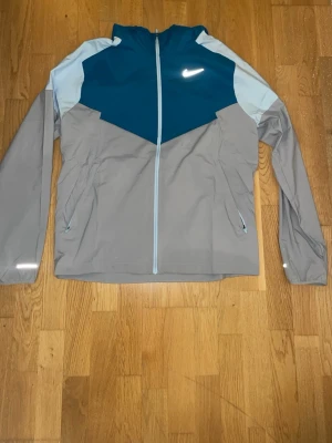 Nike vindjacka i blått och grått - Nike vindjacka med cool färgblock-design i mörkblått, ljusblått och grått. Jackan har hel dragkedja, huva och två sidofickor med dragkedja. Materialet är lätt och syntetiskt, perfekt för blåsiga dagar. Swoosh-logga på bröstet ger sportig vibe. Köpte för 1700