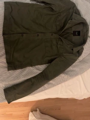 Mörkgrön overshirt från New Look - Säljer en stilren olivgrön overshirt från New Look Men. Jackan har två bröstfickor med lock, klassisk krage och knäppning framtill. Overshirten är i topp skick!