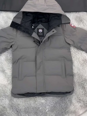 Grå pufferjacka från Canada Goose - Säljer en grå pufferjacka från Canada Goose med huva och klassisk logga på ärmen. Jackan har dragkedja, två sidofickor och quiltad design. Perfekt för kalla dagar och riktigt snygg till vinterlooken.