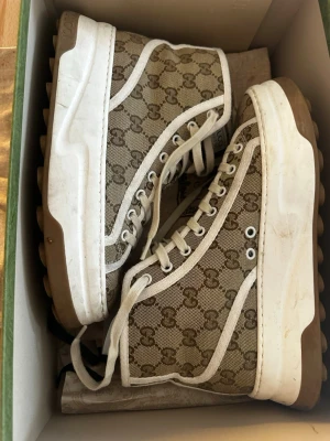 Gucci Tennis 1977 sneakers beige stl.40 - Säljer ett par Gucci Tennis 1977 high top sneakers i beige och brunt med klassiskt GG-monogram. Skorna har vita snören, kraftig vit sula och detaljer i canvas. Märkeslogga på plösen och snygg retrovibe. Perfekta för dig som vill sticka ut med lyxig streetstyle. Italienska 39, svensk stl.40