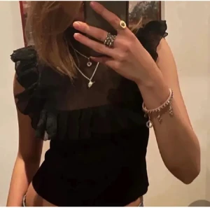 Svart volang topp med mesh - Säljer en svart topp med höghalsad design och transparent mesh upptill. Toppen har volangdetaljer längs axlar och bröst, samt är ärmlös. Materialet är en mix av mesh och syntet, vilket ger en cool och trendig look. Perfekt att styla med smycken och jeans.