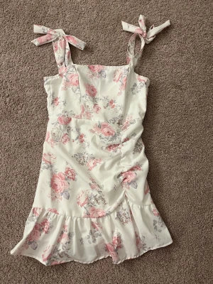 Blommig kortklänning med knytband - Söt kortklänning i ljus färg med rosa och gråa blommor. Klänningen har axelband med knyt, volangkant nedtill. Strl XS