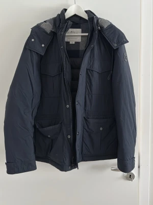 Mörkblå dunjacka från Woolrich - Dunjacka från Woolrich i mörkblå färg med huva och flera praktiska fickor framtill. Jackan har dragkedja och tryckknappar, samt ett snyggt märke på ärmen. Insidan har rutigt foder i ullblandning för extra värme. Perfekt för kyliga dagar. Inga hål, lösa sömmar eller liknande. Nypris~ 3000, mitt pris: 1300 men kan gå ner vid snabb och smidig affär.