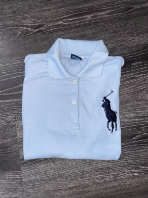 Ljusblå Ralph Lauren piké XL - Snygg ljusblå pikétröja från Ralph Lauren i storlek XL. Klassisk design med korta ärmar, krage och två knappar framtill. Stor mörkblå broderad logga på bröstet ger en clean och sportig vibe. Tillverkad i mjuk bomull som känns skön mot huden.