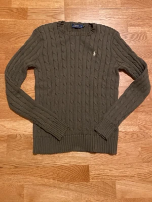 Olivgrön stickad tröja Polo Ralph Lauren - Säljer en olivgrön kabelstickad tröja från Polo Ralph Lauren. Klassisk rund halsringning, långärmad och med den ikoniska broderade loggan på bröstet. Tröjan är ribbad vid ärmslut och nederkant och har en snygg, tidlös look. Är i mycket bra skick