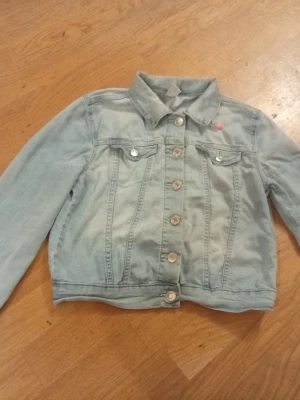 Ljusblå jeansjacka från &Denim - Säljer en ljusblå jeansjacka från &Denim i storlek 140. Jackan har klassisk krage, silverfärgade knappar och två bröstfickor. På ryggen finns handmålade detaljer med regnbåge, sol, blommor och fjärilar som gör den unik och personlig.