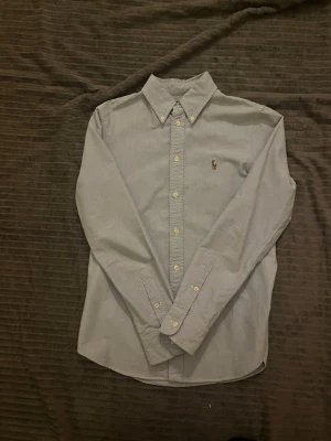 Ljusblå skjorta från Polo Ralph Lauren - Snygg ljusblå skjorta från Polo Ralph Lauren i custom fit. Klassisk button-down krage, broderad logga på bröstet och vita knappar framtill. Skjortan är långärmad och tillverkad i mjuk bomull som känns skön mot huden.