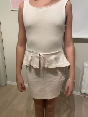 Vit peplumklänning från Zara Trafaluc - Snygg vit kortklänning från Zara Trafaluc med peplumdetalj i midjan och djup, öppen rygg. Klänningen är ärmlös och har en rundad halsringning framtill. Materialet känns mjukt och stretchigt, perfekt för dig som gillar stilrena och feminina plagg.