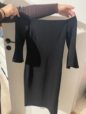 Svart offshoulder klänning med trumpetärm - Snygg svart klänning med offshoulder-design och trumpetärmar. Klänningen är figurnära och har en dragkedja i ryggen. Materialet känns mjukt och stretchigt, perfekt för dig som gillar stilrena och eleganta plagg. Passar dig som vill ha en klassisk look med en twist.