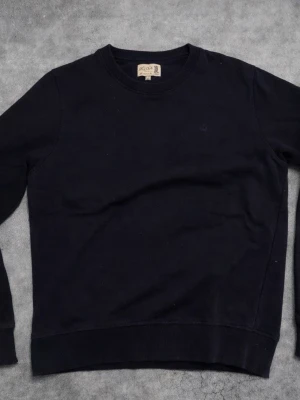 Morris - Svart sweatshirt från Morris i storlek S. Tröjan har rund hals, långa ärmar och ett diskret broderat emblem på bröstet. Tillverkad i mjuk bomull med ribbade muddar vid ärmslut och nederkant. Perfekt för chill dagar och enkel att matcha.