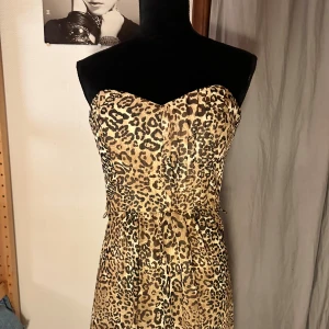 Leopardmönstrad axelbandslös långklänning - Snygg axelbandslös långklänning med leopardmönster i beige och bruna toner. Klänningen har en figurnära överdel med markerad midja och dragkedja i ryggen. Materialet är lätt och ger ett flödande fall. Perfekt för dig som vill sticka ut med ett djuriskt print.