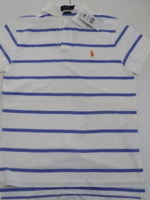 Vit och blårandig piké från Ralph Lauren - Säljer en fräsch vit pikétröja från Ralph Lauren med blå horisontella ränder och orange broderad logga på bröstet. Modellen är 'Custom Fit' och har klassisk krage samt knappar vid halsen. Perfekt för dig som gillar stilren och sportig look.