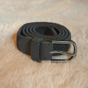 Svart flätat bälte med silverfärgat spänne - Snyggt svart bälte med flätad design och silverfärgat metallspänne. Bältet har detaljer i syntetmaterial vid ändarna och en stilren look som passar till jeans eller kostymbyxor. Flätningen ger en cool textur och gör det enkelt att justera längden.