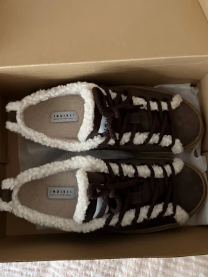 Bruna Inuikii sneakers - Säljer ett par bruna Inuikii sneakers med fluffig vit fuskpäls runt kanten och längs snörningen. Skorna har en rund tå, platt sula och är gjorda i mocka med grova bruna snören. Perfekta för dig som vill ha både stil och värme under höst och vinter. De är aldrig använda, endast testade.