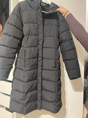 Svart lång pufferjacka från 157 - Svart pufferjacka från 157 i storlek S. Jackan är lång, har huva och är quiltad med breda kanaler. Perfekt för kalla dagar och har en stilren look. Materialet är polyester och jackan har långa ärmar samt dragkedja framtill.