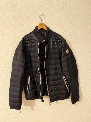  Moncler Daniel quiltad dunjacka - Svart pufferjacka från Moncler med quiltad design och coola ränder längs dragkedjorna. Jackan har hög krage, två fickor med dragkedja och Moncler-logga på ärmen. Materialet är polyester och dun, perfekt för kyliga dagar. Snygg och stilren look med långärmad passform.
