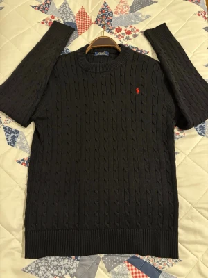 Svart kabelstickad tröja Polo Ralph Lauren - Kabelstickad svart tröja från Polo Ralph Lauren med klassisk röd logga broderad på bröstet. Tröjan har rund halsringning, ribbade muddar och är långärmad. Perfekt för dig som gillar stilrena och tidlösa plagg.Priset kan diskutera🥂