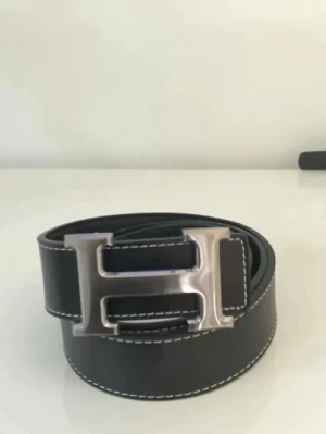 Svart skärp med H-spänne - Snyggt svart skärp med ett stort H-format metallspänne och vita kontrastsömmar längs kanterna. Perfekt accessoar för att lyfta din outfit med en stilren detalj.