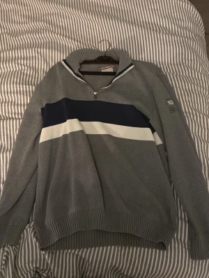 Grå half zip tröja Peak Performance XL - Säljer en grå långärmad tröja från Peak Performance i storlek XL. Tröjan har en half zip-dragkedja, ribbad krage och muddar samt breda blå och vita ränder över bröstet. Perfekt för kyligare dagar och snygg till jeans.