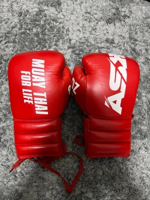 Röda Muay Thai handskar från Askari - Säljer ett par röda Muay Thai handskar från Askari med vit logga och texten 'MUAY THAI FOR LIFE'. Handskarna har snörning och är tillverkade i skinn med en cool, sportig look. Perfekta för dig som tränar kampsport och vill ha snygga handskar med bra passform.
