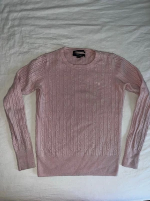 Ljusrosa stickad tröja från GANT - Säljer en ljusrosa kabelstickad tröja från GANT med rund hals och diskret logga broderad på bröstet. Tröjan har långärmad passform och breda ribbade muddar vid ärmslut och nederkant. Perfekt för dig som gillar klassisk och stilren look. Den är storlek M men ganska tajt. Skulle säga att den är storlek S om man vill att det ska vara lite mer släpt. 