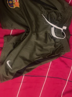 Gröna Nike FC Barcelona shorts L - Säljer ett par gröna FC Barcelona shorts från Nike i storlek L. De har elastisk midja med vit snörning och är gjorda i lätt polyester. Perfekta för fotboll eller träning, med klubbmärke och Nike-logga på framsidan. Oanvända.
