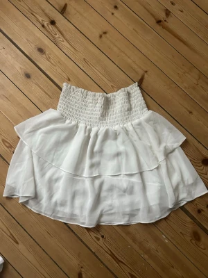 Vit volangkjol från Made by Chelsea - Superfin vit kor tkjol från Made by Chelsea i storlek S. Kjolen har smock i midjan och dubbla lager med volanger som ger en flowy och luftig känsla. Materialet känns lätt och skönt mot huden.