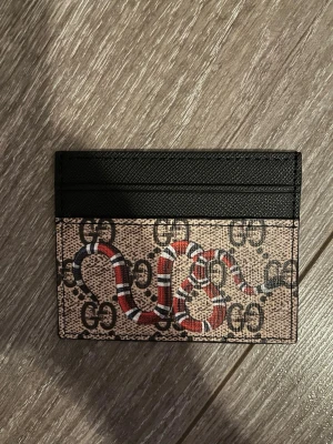 Korthållare i beige med orm  - Säljer en snygg korthållare med klassiskt GG-mönster och en cool röd, vit och svart orm på framsidan. Plånboken är i beige och svart med flera kortfack och är tillverkad i tåligt skinn. Perfekt för dig som vill ha något unikt och trendigt i fickan.