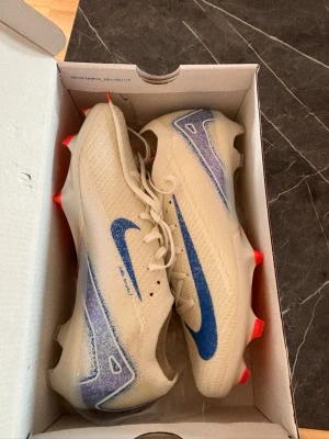 Nike Zoom Vapor 16 Elite FG fotbollsskor - Nike Zoom Vapor 16 Elite FG fotbollsskor i vit med blå swoosh och lila detaljer. Skorna har snörning, platt sula med röda dobbar och är tillverkade i syntetmaterial för grym komfort och grepp på planen. Perfekt för dig som vill ha max prestanda på matchen.