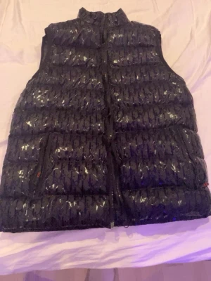Svart dunväst från Christian Dior - Säljer en svart dunväst från Christian Dior i storlek M. Västen har ett glansigt material med ett tydligt Dior-mönster över hela ytan, två fickor med dragkedja och hög krage. Perfekt för att addera lite lyx till din outfit.