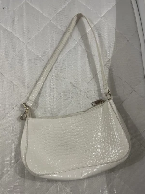 Vit axelväska med krokodilmönster - Säljer en snygg vit axelväska med glansig yta och krokodilmönster. Väskan har en smal axelrem och stängs med dragkedja upptill. Perfekt för dig som gillar stilrena accessoarer med lite edge.